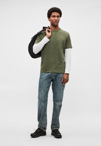 Oliivinvihreä puuvillainen t-paita valkoisen pitkähihnaisen paidan päällä, yhdistetty sinisiin farkkuihin ja mustiin tennareihin, mukana musta reppu.