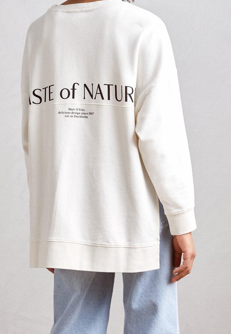 Personne portant un sweat-shirt écru avec un texte noir "TASTE OF NATUR" et un texte plus petit au dos, associé à un jean bleu clair.