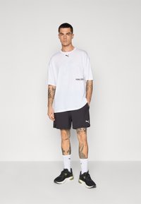 Hvit oversized t-skjorte med rund hals, med teksten "PUMA CREW" på siden, kombinert med mørke shorts og svarte joggesko med grønne detaljer.