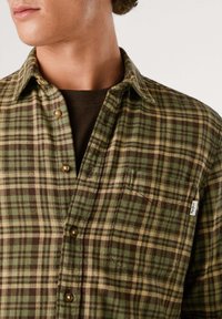 Grønn og brun rutete flanellskjorte med brystlomme, knapper på kragen og beige knapper. Har en myk tekstur og synlige sømmer.