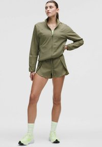 Olivgrön träningsklädesset med strukturerad zip-jacka och matchande shorts. Har en figurnära design och lagerdetaljer. Neonskor och strumpor.