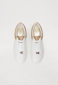 Baltas sporta apavi ar gludu ādas virsmu, zeltainiem akcentiem un teksturētu bežīgu aizmuguri. Orange šņores un logotipa detaļa priekšā.