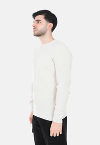 Maglione a maglia beige chiaro con texture a costine e collo rotondo, abbinato a pantaloni cargo neri. Vista di profilo.