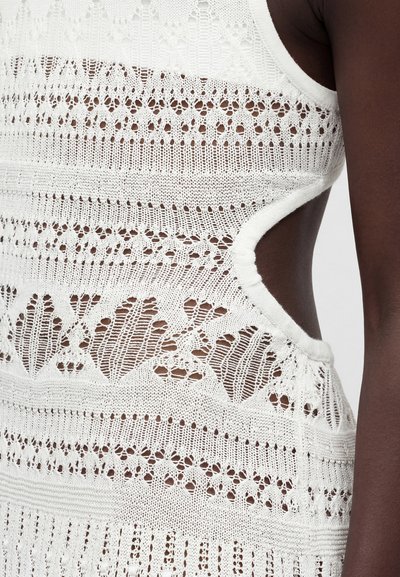 Κοντινό πλάνο ενός ατόμου με σκουρόχρωμο δέρμα που φοράει λευκό πλεκτό φόρεμα με δαντελένια σχέδια και πλευρικό άνοιγμα που δείχνει τον δέρμα.