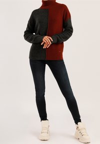 Dunkelgrauer und rostfarbener Turtleneck-Pullover mit gesplittetem Design, aus strukturiertem Strickmaterial. Kombiniert mit dunklen Jeans und weißen Stiefeln.