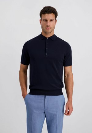 Ein Mann steht in dunklem marineblauem Kurzarm-Poloshirt und hellblauer Hose vor einem schlichten hellen Hintergrund und schaut nach vorne.