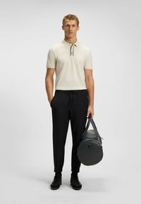Beige Polohemd mit einem schwarz-weißen Kragen, schwarze Sweatpants und schwarze Sportschuhe. Hält eine graue Reisetasche mit Griffen.