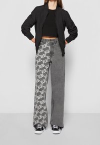 Donna che indossa jeans grigi a due tonalità con stampa bianca su una gamba, top corto nero, giacca bomber nera e sneakers bianco e nere.