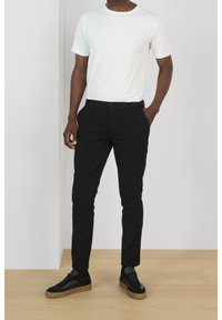 Bílý tričko s krátkým rukávem v kombinaci s těsnými černými kalhotami a černými sneakers s hnědými podešvemi, umístěné na neutrálním pozadí.
