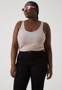 Mujer con gafas de sol rosas, pendientes de aro, un top sin mangas a rayas de múltiples colores y pantalones negros, mirando hacia un lado sobre un fondo liso.