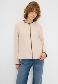 Giacca trapuntata beige con collo rotondo, bordatura marrone e bottoni dorati. Abbinata a jeans a gamba ampia blu. Tessuto liscio.