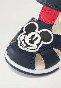 Tamsiai mėlyni odiniai sandalai su atvira dizaino koncepcija, turintys baltą Mickey Mouse emblemos ir raudoną lipnų dirželį. Minkštas vidus ir tekstūruotas padas.