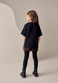 Czarny, oversize t-shirt, szorty w kratkę i czarne legginsy. Dziewczyna w czarnych botkach sięgających do kostki; widok z tyłu na neutralnym tle.