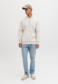 Jaunas vyras dėvi smėlio spalvos pusiau užtraukiamą džemperį, šviesiai mėlynas džinsus, smėlio spalvos sportbačius ir smėlio spalvos kepurę, stovįs priešais paprastą baltą foną.