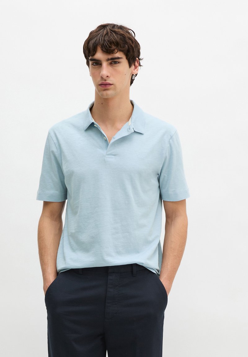 Jeune homme portant un polo à manches courtes bleu clair et un pantalon sombre, debout les mains dans les poches devant un fond blanc uni.