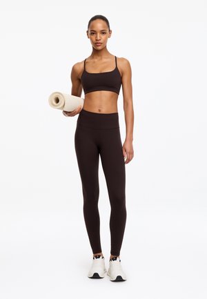 Jeune femme en tenue de sport noire et baskets blanches tenant un tapis de yoga roulé, debout devant un fond blanc uni.