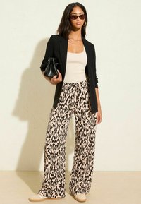 Kvinna klädd i svart blazer, krämfärgad linne, vida djurmönstrade byxor, beige sneakers, solglasögon och bärande på en svart handväska mot en enfärgad bakgrund.