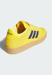 Baskets Adidas jaunes avec des rayures bleues, panneaux en daim et en cuir, semelle beige et lacets, montrées sous un angle arrière.