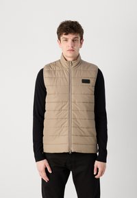 Μπεζ γιλέκο με κάθετη ραφή, ψηλό γιακά, μπροστινή φερμουάρ και μαύρη επιγραφή λογότυπου στο αριστερό στήθος. Μαύρο μακρυμάνικο πουκάμισο από κάτω.