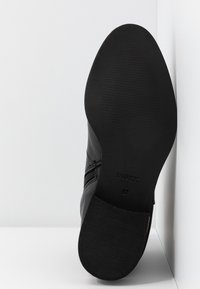 Chaussure noire avec une tige en cuir texturé, semelle en caoutchouc et bord festonné. Présente un logo clairement imprimé sur la semelle ; taille 37.