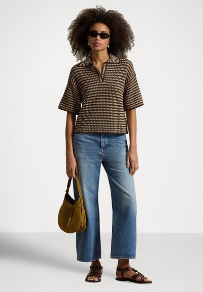 Ženska s kodrastimi lasmi, ki nosi temna sončna očala, rjav pulover s črtami in kratkimi rokavi, modre kavbojke, drži gorčično torbico in ima rjave sandale.