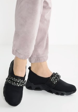 Chaussures noires à enfiler avec embellissement en perles, dessus en synthétique, semelle texturée et design arrondi. Portées avec un pantalon léger rose à revers.