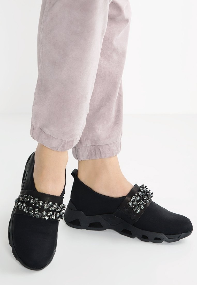 Chaussures noires à enfiler avec embellissement en perles, dessus en synthétique, semelle texturée et design arrondi. Portées avec un pantalon léger rose à revers.