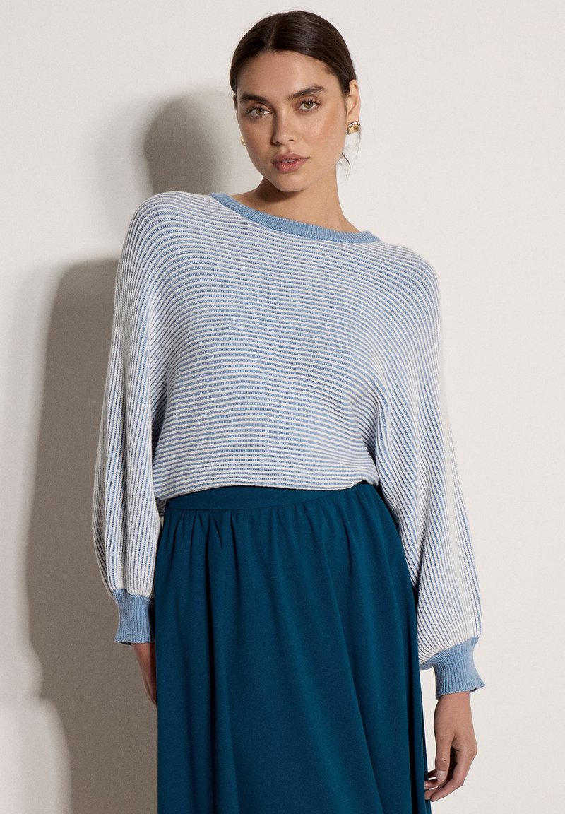 Femme portant un pull rayé bleu clair et blanc avec des manches bouffantes et une jupe taille haute bleu foncé, debout contre un mur uni.