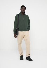 Grøn kvart-zip trøje i blødt stof, beige bukser og sorte sko. Modellen holder en sort skuldertaske. Enkel design med minimale detaljer.
