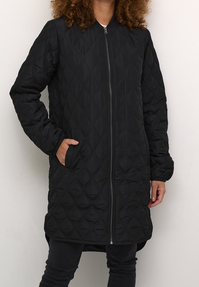 Femme portant une longue veste matelassée noire zippée avec des poches latérales et un pantalon noir, debout contre un mur de couleur claire.