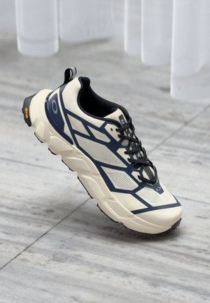 Chaussure de course légère avec une tige crème, des accents bleu marine, un mesh texturé et une semelle rembourrée ; dotée d'un talon en caoutchouc noir et d'un logo.