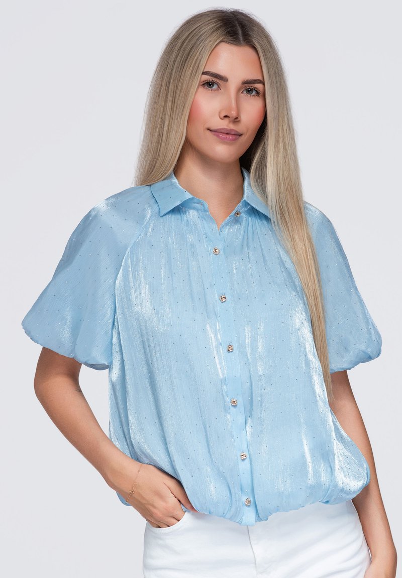 Femme aux longs cheveux blonds lisses portant un chemisier court brillant bleu clair et un pantalon blanc, avec une main dans la poche.