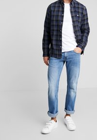 Hombre con una camisa de cuadros azul y negro sobre una camiseta blanca, jeans claros con puños y zapatillas blancas, con una mano en el bolsillo.