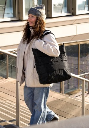 Göndör hajú nő szürke kötött sapkában és fehér kabátban, nagy fekete táskát visel, a szabadban lépcsőn áll.