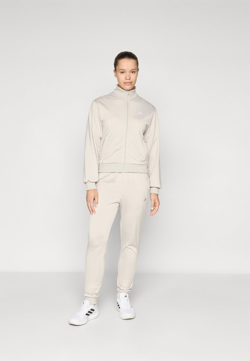 Lys beige treningsdress laget av myk stoff, med en jakke med glidelås og ribbede mansjetter, samt matchende bukser. Tre-stripe detaljer og logo.