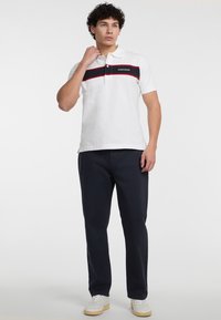 Camisa polo blanca con una franja horizontal negra y roja, que cuenta con un cuello, dos botones y el logo "GUESS JEANS". Combinada con pantalones azul marino.