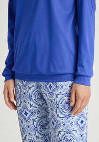 Maglietta a maniche lunghe blu abbinata a legging con motivo floreale in tonalità di blu e verde su sfondo bianco.