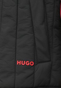 Giacca a vento nera imbottita con texture trapuntata, con logo rosso "HUGO" sul lato inferiore e una tasca laterale senza componenti hardware visibili.