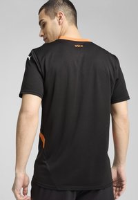 Camiseta deportiva negra con cuello y acentos laterales naranjas, manga corta y tela transpirable, con un logo cerca del cuello.
