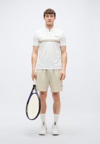 Λευκό αθλητικό πουκάμισο με φερμουάρ στο γιακά και καφέ ρίγες, συνδυασμένο με μπεζ σορτς. Το μοντέλο κρατά ρακέτα τένις, φοράει λευκά αθλητικά παπούτσια και κάλτσες.