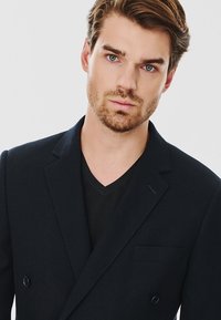 Granatowa, podwójnie zapinana marynarka o strukturalnym fasonie. Posiada klapę typu notch, jedną kieszeń oraz teksturowany materiał. Nosi się ją na czarnej koszulce z dekoltem w serek.