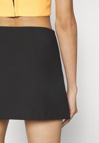 Mini-jupe noire à texture lisse, avec des coutures visibles le long des coutures. Le design est simple et moulant.