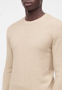 Gros plan d'une personne portant un pull en maille beige texturé avec un col rond, montrant le haut du torse et le cou.