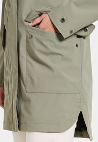 Olivgrön jacka med ett slätt, vattenavvisande tyg. Har en sidoficka för händerna och tryckknappar, samt en delad nederkant för ökad rörelsefrihet.