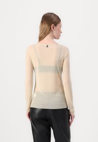 Beige langermet topp i gjennomsiktig stoff, med logo detalj på ryggen og tettsittende design, kombinert med svarte høyt midje bukser.