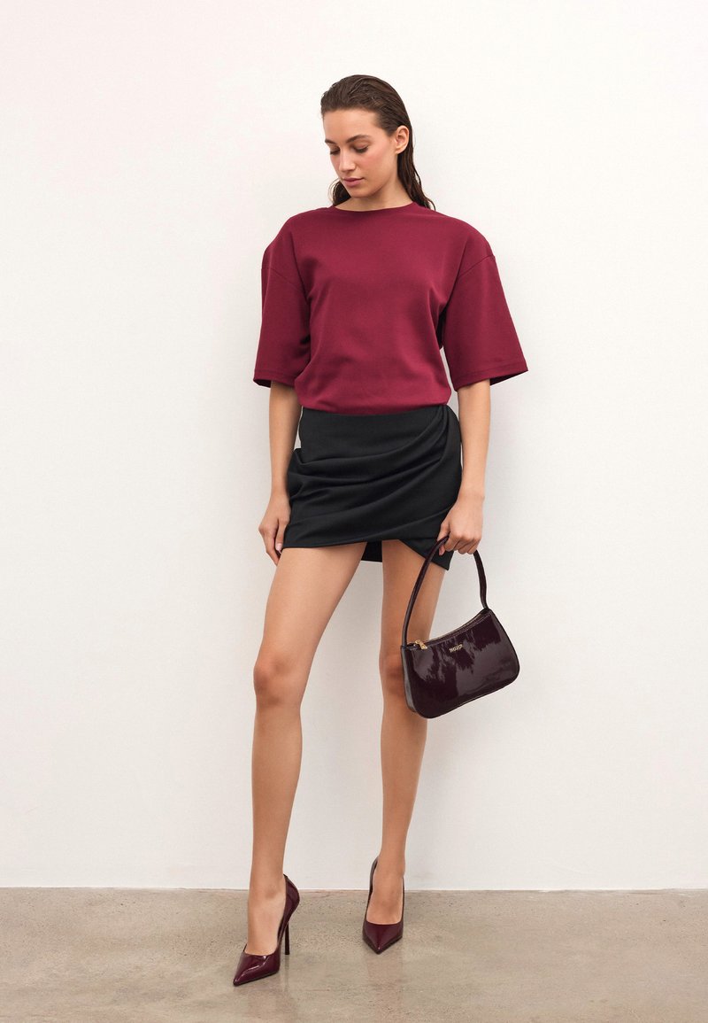 Burgundijas krāsas īsām piedurknēm topam, kas apvienots ar melnu mini svārku. Modelis tur spīdīgu burgundijas krāsas rokassomu un valkā atbilstošus augstpapēžu apavus.