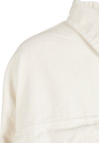 Gros plan sur l'épaule et le col d'une chemise en velours côtelé blanc, montrant la texture côtelée verticale et les détails des coutures.