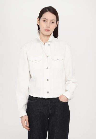 Chaqueta de mezclilla blanca cortada con cuello alzado, dos bolsillos en el pecho, botones plateados y manga larga, combinada con jeans negros.