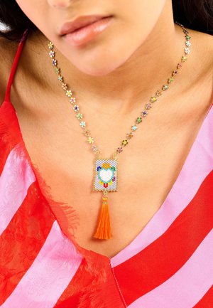 Bunte, florale Perlenkette mit einem rechteckigen Anhänger, der ein Herzmotiv und eine orangefarbene Quaste zeigt, vor Haut und Stoff.