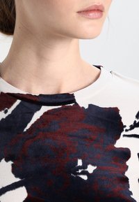 Zbliżenie jasnego ubrania z kwiatowym wzorem w ciemnoczerwonych i granatowych odcieniach, okrągłym dekoltem oraz miękką strukturą materiału.